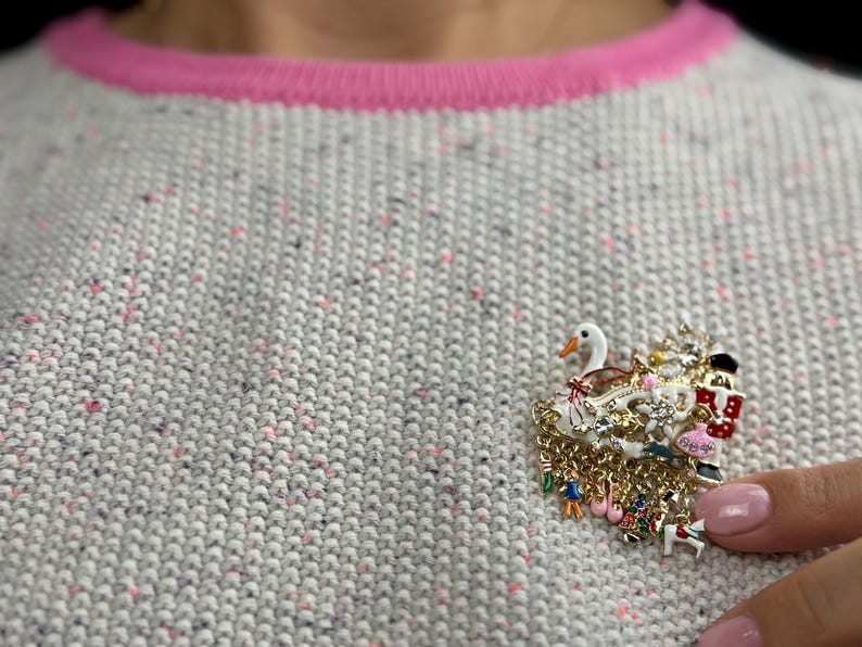 May include: A decorative brooch featuring a swan, Christmas tree, and other festive elements. The brooch is made of gold-toned metal with colorful enamel details and dangling charms. It is pinned to a gray sweater with a pink neckline.