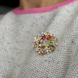 May include: A gold-toned Christmas wreath brooch with red, green, and white accents. The brooch features miniature figures and a red bow. The brooch is pinned to a gray sweater with pink trim.
