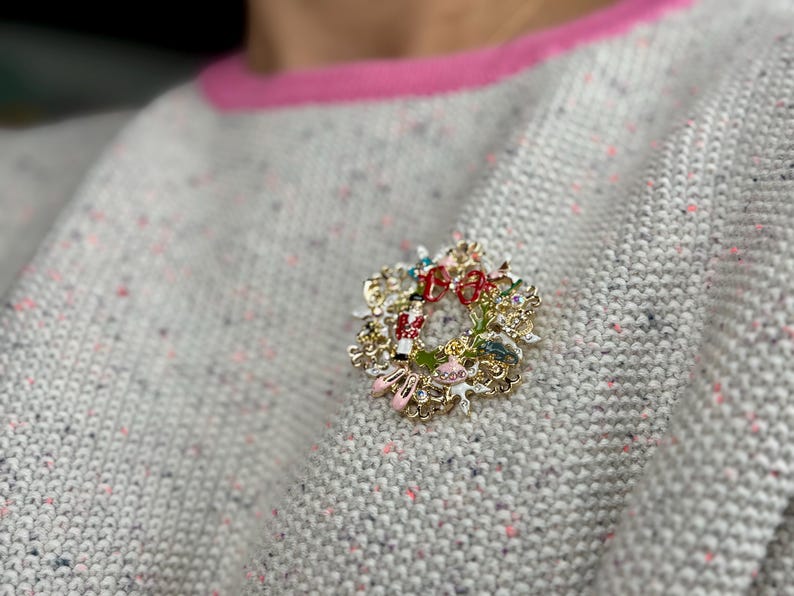 May include: A gold-toned Christmas wreath brooch with red, green, and white accents. The brooch features miniature figures and a red bow. The brooch is pinned to a gray sweater with pink trim.