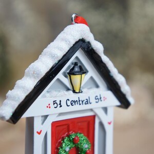 May include: White and black miniature house ornament with a red door and a wreath. The house has a snowy roof and a street sign that reads "51 Central St."