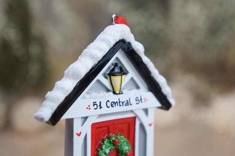 May include: White and black miniature house ornament with a red door and a wreath. The house has a snowy roof and a street sign that reads "51 Central St."