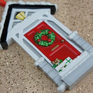 May include: A white and gray Christmas ornament shaped like a house with a red door and a green wreath. The door has a small silver handle and a white trim. The house has a white roof with a black trim and a small window with a yellow light.