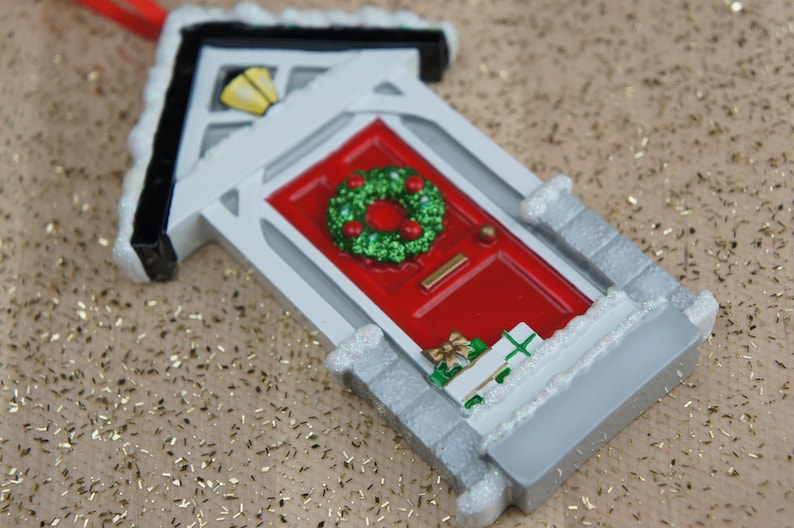 May include: A white and gray Christmas ornament shaped like a house with a red door and a green wreath. The door has a small silver handle and a white trim. The house has a white roof with a black trim and a small window with a yellow light.