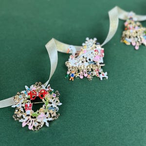 May include: Three Christmas-themed brooches with gold-tone metal and colorful enamel details. One is a wreath with a red bow, another is a Christmas tree, and the third is a snowman. Each brooch is attached to a cream-colored ribbon.