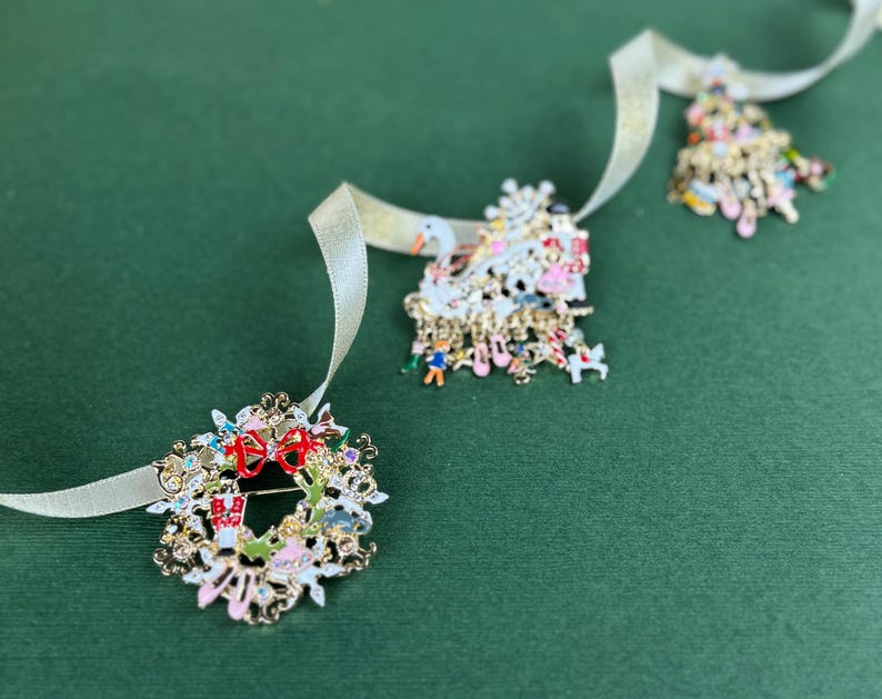 May include: Three Christmas-themed brooches with gold-tone metal and colorful enamel details. One is a wreath with a red bow, another is a Christmas tree, and the third is a snowman. Each brooch is attached to a cream-colored ribbon.