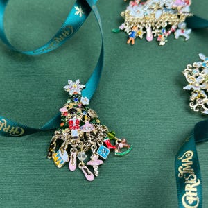 May include: Christmas tree-shaped brooch with dangling charms, including ballet slippers and a rocking horse. The brooch is gold-toned with colorful enamel details. A teal ribbon with "CHRISTMAS" text is in the image.