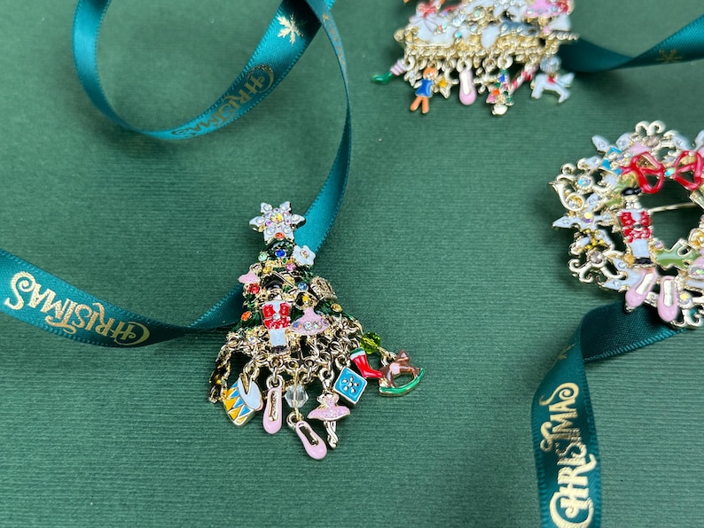 May include: Christmas tree-shaped brooch with dangling charms, including ballet slippers and a rocking horse. The brooch is gold-toned with colorful enamel details. A teal ribbon with "CHRISTMAS" text is in the image.
