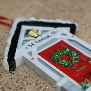 May include: White ceramic house ornament with a red door and a green wreath. The house has a black roof and a white sign that reads "51 Central St." and "2023".