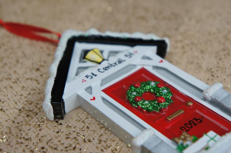 May include: White ceramic house ornament with a red door and a green wreath. The house has a black roof and a white sign that reads "51 Central St." and "2023".