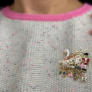 May include: A gold-toned brooch with a swan, Christmas-themed elements, and dangling charms. The brooch is set against a gray sweater with pink trim. The brooch features a swan, a nutcracker, and other festive charms.