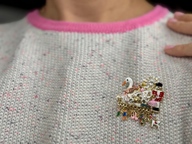 May include: A gold-toned brooch with a swan, Christmas-themed elements, and dangling charms. The brooch is set against a gray sweater with pink trim. The brooch features a swan, a nutcracker, and other festive charms.