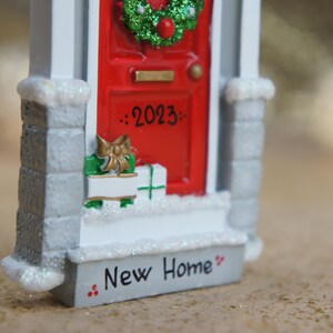 May include: A white and gray miniature house ornament with a red door, a green wreath, and the text "2023" and "New Home" on the front.
