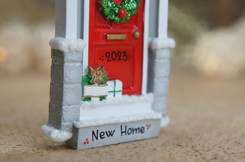 May include: A white and gray miniature house ornament with a red door, a green wreath, and the text "2023" and "New Home" on the front.