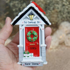 May include: White and red house-shaped Christmas ornament with a red door, a wreath, and the text "51 Central St." and "New Home".