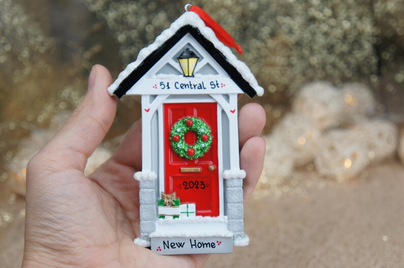 May include: White and red house-shaped Christmas ornament with a red door, a wreath, and the text "51 Central St." and "New Home".