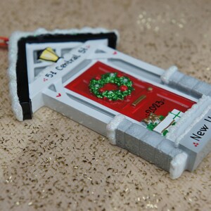 May include: A white and gray house-shaped Christmas ornament with a red door, a green wreath, and the text "54 Central St." and "New Home".