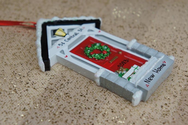 May include: A white and gray house-shaped Christmas ornament with a red door, a green wreath, and the text "54 Central St." and "New Home".