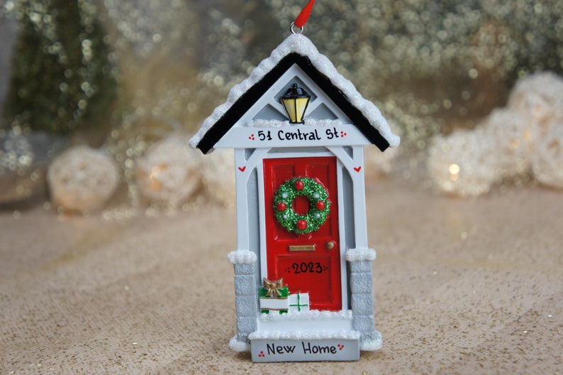 May include: A white and gray house-shaped Christmas ornament with a red door, a green wreath, and the text "51 Central St." and "New Home".