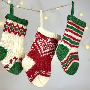 May include: Three small, hand-knitted Christmas stockings hanging from a string. The left stocking is cream with green and red patterns. The center stocking is red with a white heart design. The right stocking features horizontal red and green stripes.