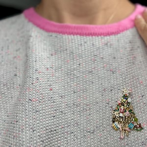 May include: A gray sweater with a pink trim and a Christmas tree charm. The sweater has a speckled pattern with pink and black flecks. The charm is a detailed Christmas tree with various ornaments.