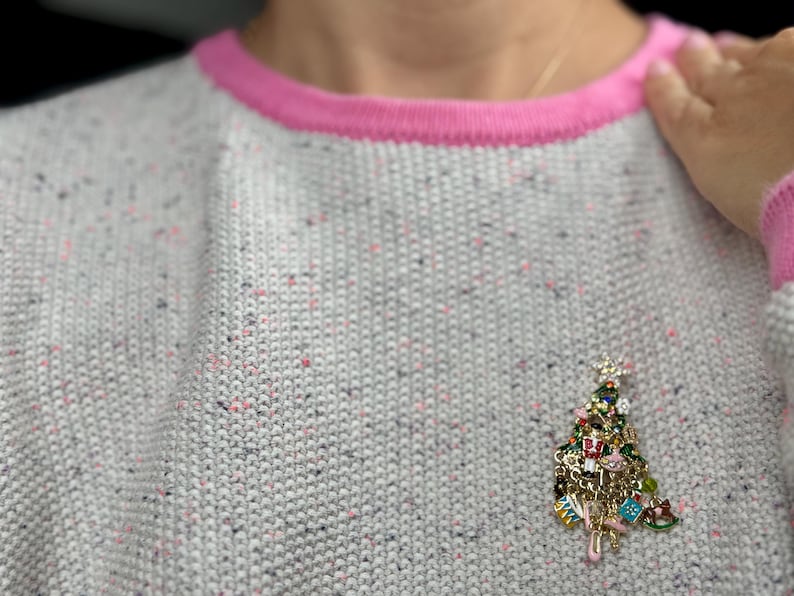 May include: A gray sweater with a pink trim and a Christmas tree charm. The sweater has a speckled pattern with pink and black flecks. The charm is a detailed Christmas tree with various ornaments.