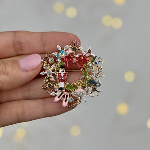 May include: A festive Christmas wreath brooch with a gold-tone finish. The brooch features a red bow, a toy soldier, ballet slippers, and other holiday-themed embellishments. The wreath is adorned with colorful enamel and sparkling crystals.