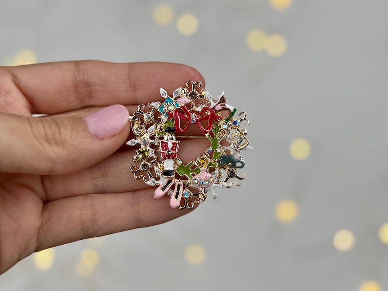 May include: A festive Christmas wreath brooch with a gold-tone finish. The brooch features a red bow, a toy soldier, ballet slippers, and other holiday-themed embellishments. The wreath is adorned with colorful enamel and sparkling crystals.