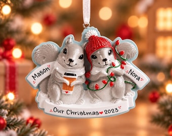 May include: A hanging Christmas ornament featuring two gray squirrels. One squirrel wears a red knit hat and holds a string of red lights, while the other holds a coffee cup. The ornament includes the names Mason and Nora and the text Our Christmas 2023.