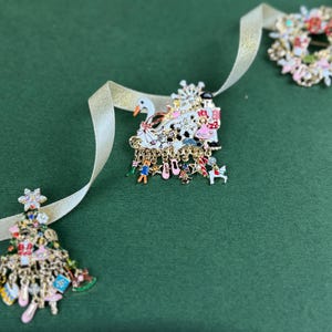May include: A collection of Christmas-themed brooches and pins. The brooches include a Christmas tree, a swan, and a wreath, all with colorful enamel details and dangling charms. A gold ribbon weaves through the scene against a green background.