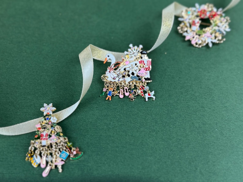 May include: A collection of Christmas-themed brooches and pins. The brooches include a Christmas tree, a swan, and a wreath, all with colorful enamel details and dangling charms. A gold ribbon weaves through the scene against a green background.