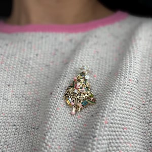 May include: A gold-toned Christmas tree brooch with colorful enamel and charm details. The brooch is pinned to a light gray sweater with pink trim. The tree is adorned with various charms and colorful accents.