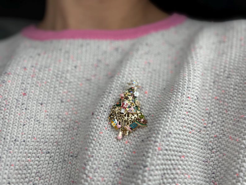 May include: A gold-toned Christmas tree brooch with colorful enamel and charm details. The brooch is pinned to a light gray sweater with pink trim. The tree is adorned with various charms and colorful accents.