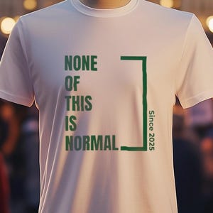 May include: White t-shirt with the text "NONE OF THIS IS NORMAL" and "Since 2015" printed on it. The shirt is on a mannequin in front of a blurred background of a crowd of people.