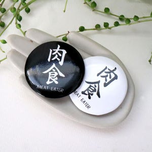 May include: Two round buttons, one black and one white, each with Asian characters and the English text "MEAT EATER". The buttons are resting on a gray, hand-shaped dish, with green plant stems and small round leaves in the background.