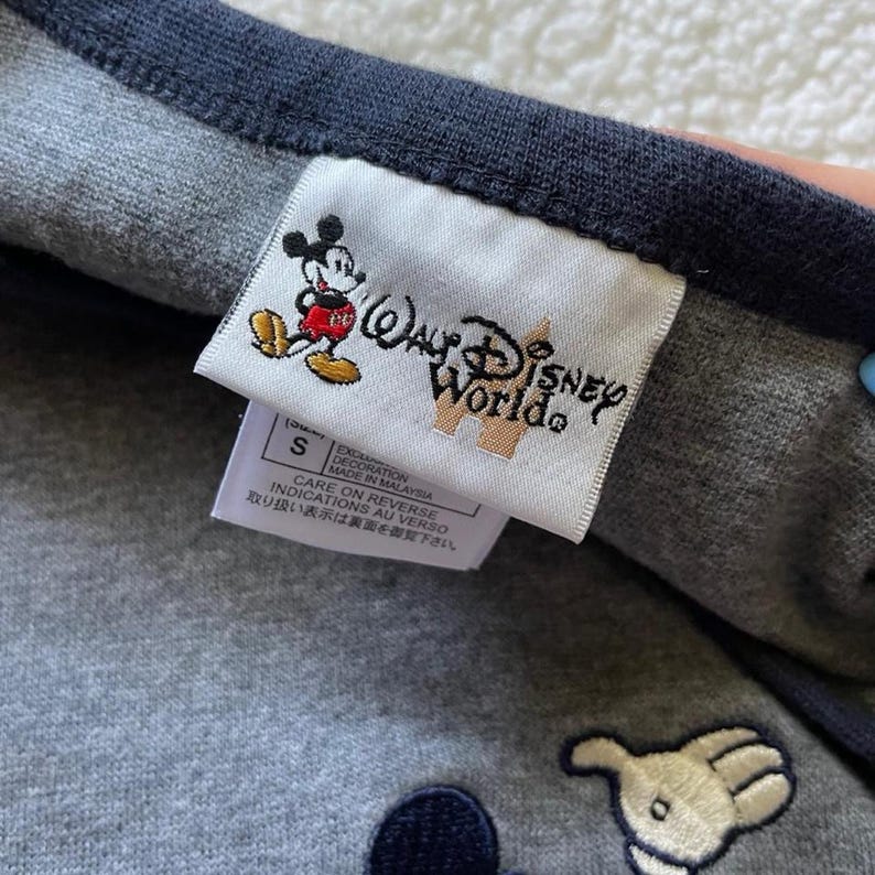 May include: A grey garment with a dark blue trim featuring a white fabric tag. The tag displays a graphic of Mickey Mouse and the text Walt Disney World. A secondary white tag below indicates size S, Made in Malaysia, and care instructions.