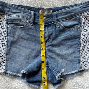 May include: Light blue denim shorts with a frayed hem and white lace side panels. The shorts measure approximately 10 inches in length. The shorts have a button and zipper closure and front and back pockets.