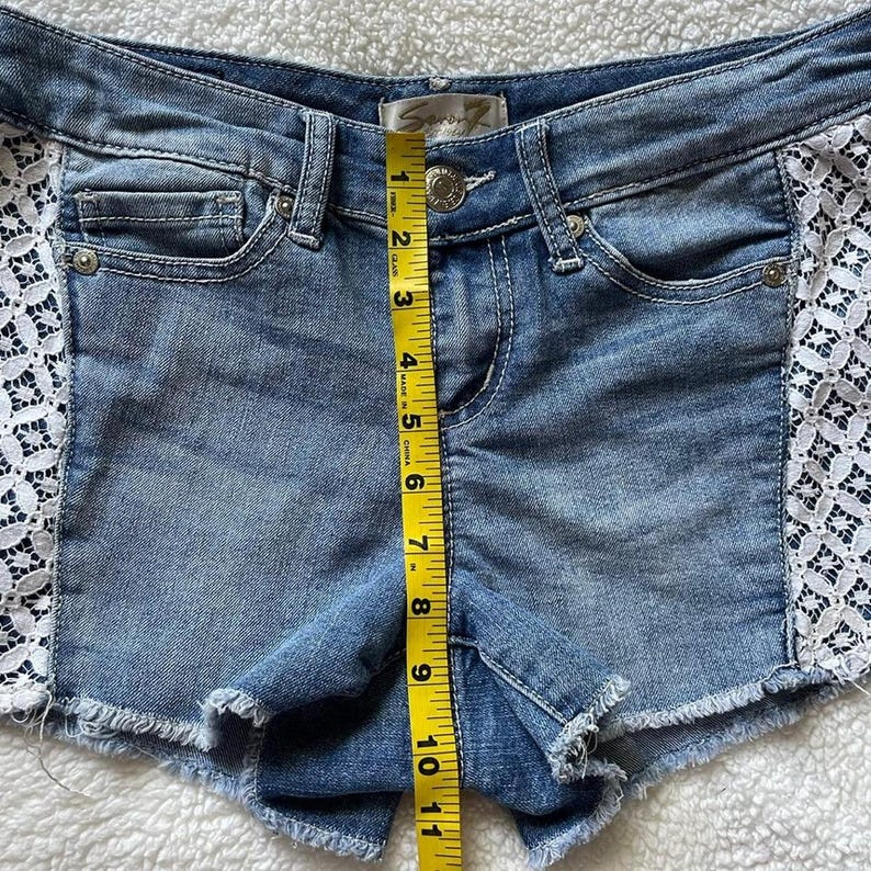 May include: Light blue denim shorts with a frayed hem and white lace side panels. The shorts measure approximately 10 inches in length. The shorts have a button and zipper closure and front and back pockets.