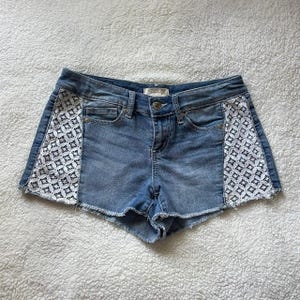 May include: Light blue denim shorts with white lace panels on the sides. The shorts have a classic five-pocket design, a button and zipper closure, and a raw hem. The shorts are made of a light wash denim.