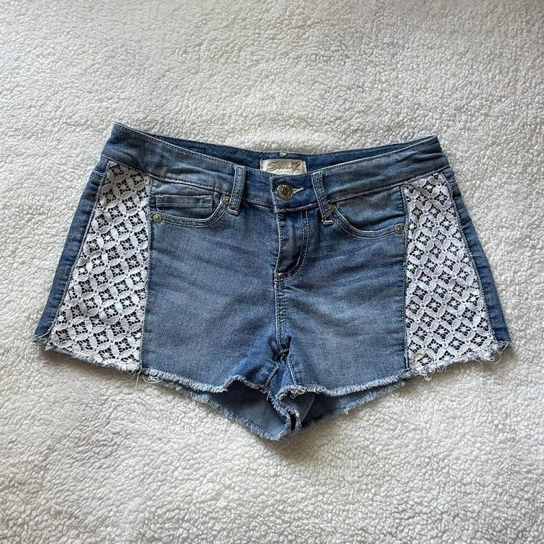 May include: Light blue denim shorts with white lace panels on the sides. The shorts have a classic five-pocket design, a button and zipper closure, and a raw hem. The shorts are made of a light wash denim.