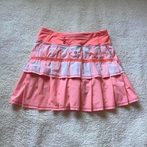 Può includere: Una gonna sportiva color rosa corallo caratterizzata da un design plissettato a balze con una tasca orizzontale con cerniera in vita. Il capo presenta un piccolo logo circolare centrato sul pannello superiore e una struttura a strati con cuciture a vista su tutto il tessuto.