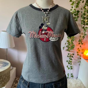 May include: A heather grey short-sleeve t-shirt with a dark grey crew neckline. The front features a graphic of Mickey Mouse inside a red circle with the text Mickey Mouse Since 1928. A silver butterfly pendant on a beaded necklace is worn over the shirt.