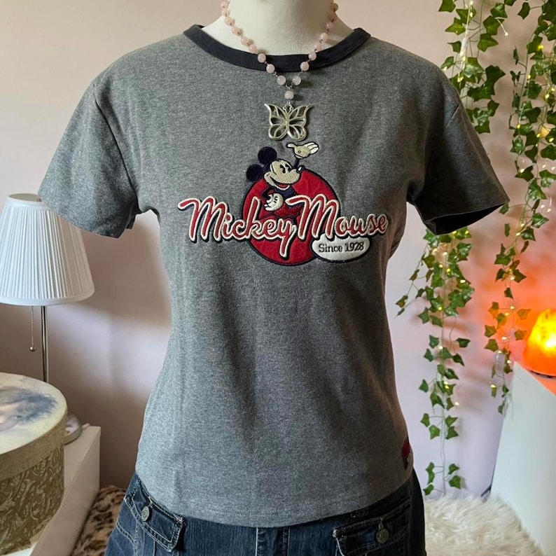 May include: A heather grey short-sleeve t-shirt with a dark grey crew neckline. The front features a graphic of Mickey Mouse inside a red circle with the text Mickey Mouse Since 1928. A silver butterfly pendant on a beaded necklace is worn over the shirt.