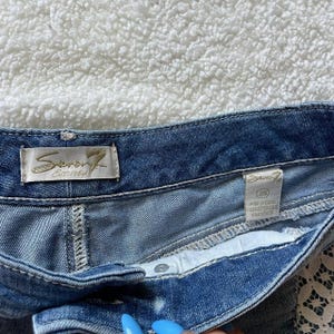May include: Close-up of blue denim shorts. The shorts have a button closure and a white interior. The brand label "Seven7 Est. 1969" is visible. The shorts are size 28.