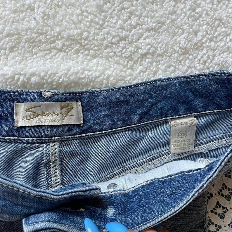 May include: Close-up of blue denim shorts. The shorts have a button closure and a white interior. The brand label "Seven7 Est. 1969" is visible. The shorts are size 28.