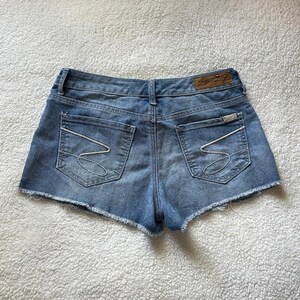 May include: Light blue denim shorts with a raw hem. The shorts have two back pockets with white stitching and a small leather patch with the brand name. The back of the shorts are visible.