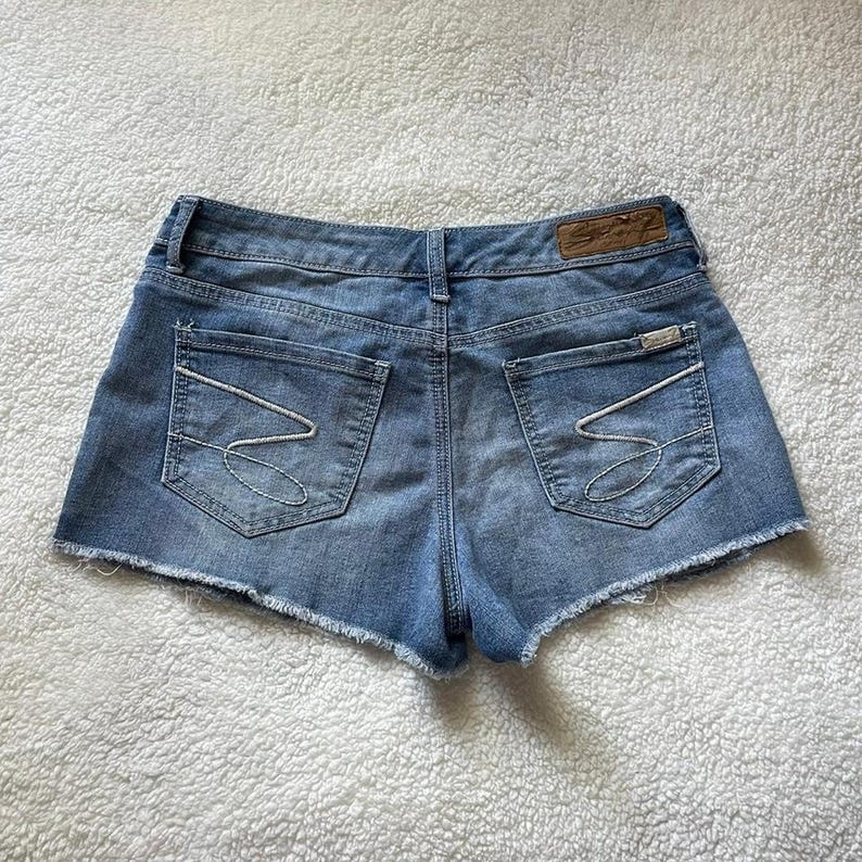 May include: Light blue denim shorts with a raw hem. The shorts have two back pockets with white stitching and a small leather patch with the brand name. The back of the shorts are visible.