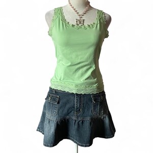 May include: A light green sleeveless tank top with a scalloped lace trim along the neckline and hem, paired with a dark blue denim mini skirt featuring two front flap pockets and a ruffled bottom edge, accessorised with a pink beaded butterfly necklace.