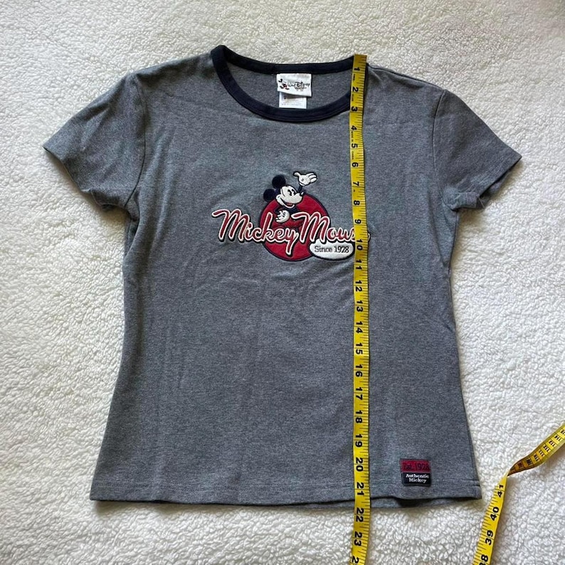 May include: A grey short-sleeved t-shirt with a dark blue crew neckline. The front features a Mickey Mouse graphic with the text "Mickey Mouse Since 1928". A yellow measuring tape placed vertically shows a length of approximately 58.5 cm.