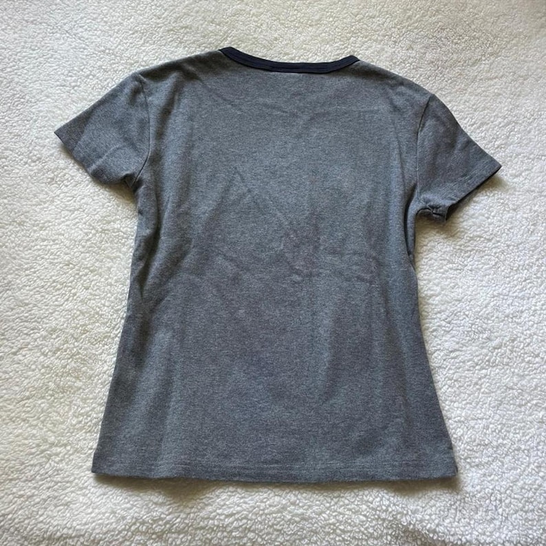 May include: A short-sleeved t-shirt in a heathered grey fabric featuring a solid dark navy blue crew neckline trim. The garment is displayed flat, showcasing a simple, plain design with a standard cut, short sleeves, and a straight hemline.