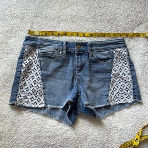 May include: Light blue denim shorts with white lace panels on the sides. The shorts have a raw hem and classic five-pocket styling. The shorts measure approximately 18 inches across.